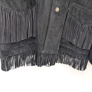 May include: Close-up of a black suede jacket with fringe detailing. The jacket has fringe along the pockets and hemline, and a button closure. The suede has a soft, textured appearance.