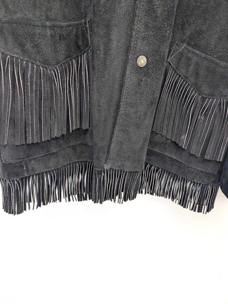 May include: Close-up of a black suede jacket with fringe detailing. The jacket has fringe along the pockets and hemline, and a button closure. The suede has a soft, textured appearance.