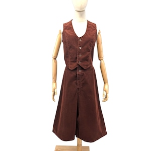 Puede incluir: Un conjunto vintage marrón de pana, compuesto por un chaleco y una falda. El chaleco tiene cuello en V y cierre de botones. La falda es hasta la rodilla y tiene cierre de botones. El conjunto se exhibe en un maniquí de madera.