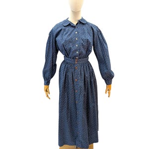 Puede incluir: Vestido vintage azul oscuro con estampado floral blanco, exhibido en un maniquí. El vestido tiene mangas largas, una botonadura frontal y una cintura entallada. La falda es larga y el vestido está hecho de una tela estampada.