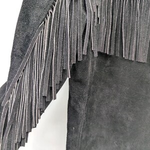 May include: Close-up of a black suede jacket with long fringe detailing along the sleeves and upper chest. The fringe is made of the same material as the jacket. The jacket has a classic, western-inspired design.