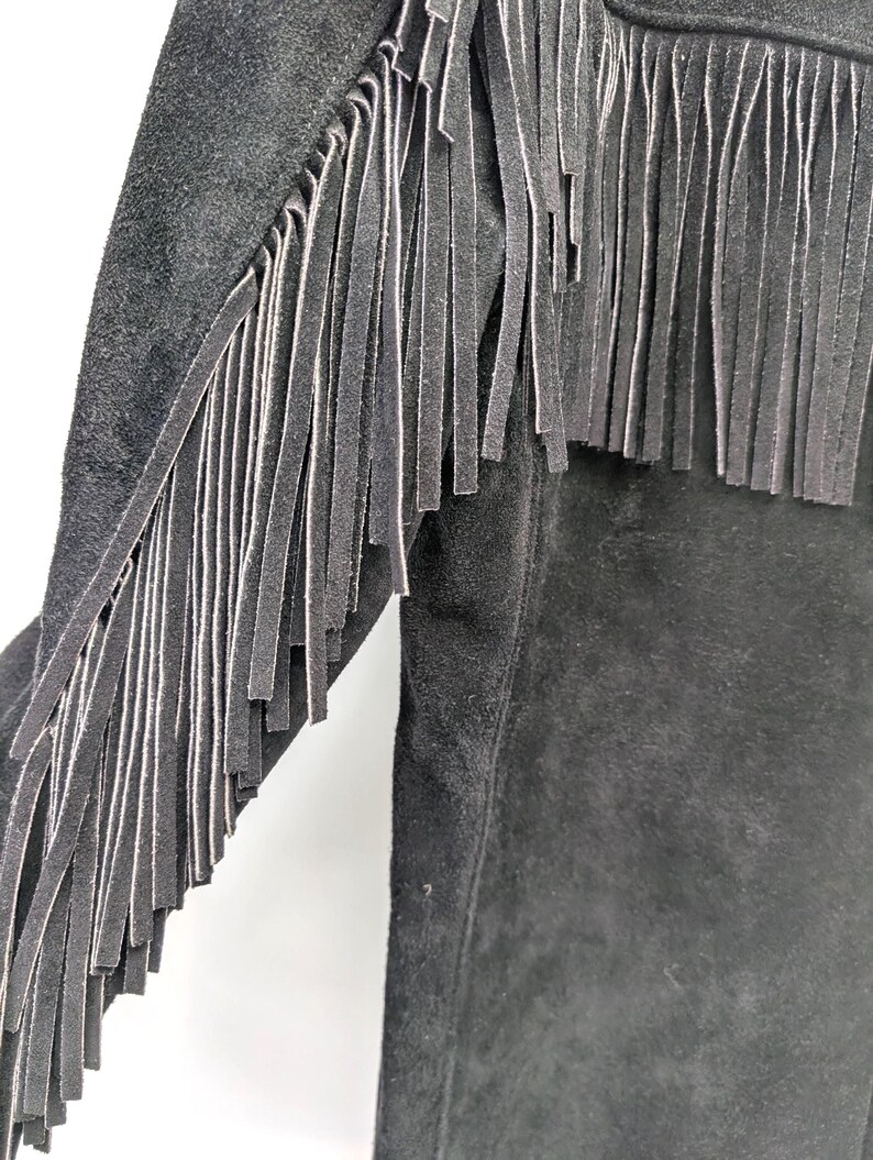 May include: Close-up of a black suede jacket with long fringe detailing along the sleeves and upper chest. The fringe is made of the same material as the jacket. The jacket has a classic, western-inspired design.
