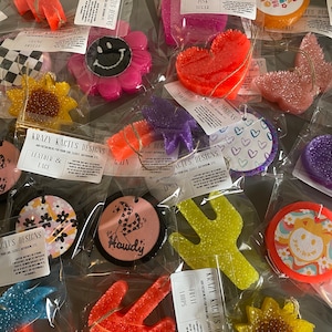 May include: Assortment of colorful car air fresheners in various shapes, including a smiley face, a heart, a sunflower, a cactus, and a cowboy boot. The air fresheners are made of a sparkly, glittery material and are packaged in clear plastic bags. The text "Krazy Kactus Designs" is visible on several of the bags.