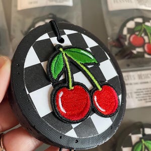 May include: A black and white checkered car air freshener with a red cherry embroidered design. The air freshener has a black string for hanging.