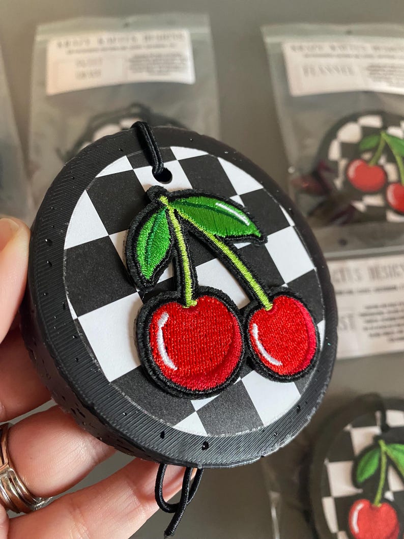 May include: A black and white checkered car air freshener with a red cherry embroidered design. The air freshener has a black string for hanging.