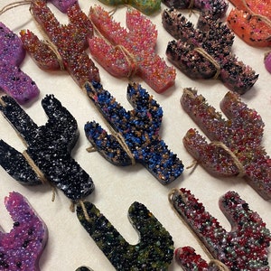 May include: Assortment of colorful cactus-shaped car air fresheners. Each cactus is made with a variety of colors and textures, including glitter and beads. The fresheners are tied with twine.