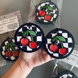 May include: Three black and white checkered car air fresheners with red cherries and green leaves. The text "KACTUS DESIGNS" and "SLEET GRACE" are visible on the packaging.