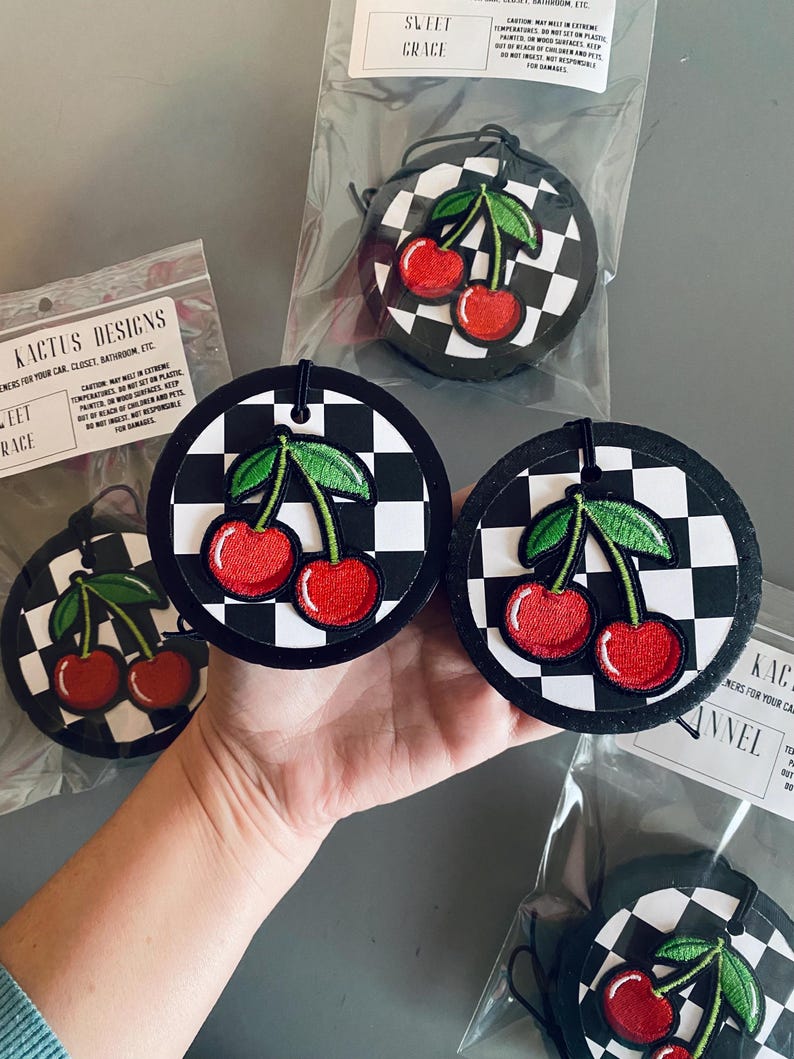 May include: Three black and white checkered car air fresheners with red cherries and green leaves. The text "KACTUS DESIGNS" and "SLEET GRACE" are visible on the packaging.