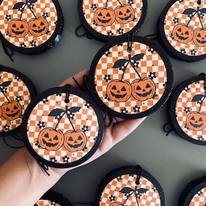 May include: Round Halloween-themed decorations featuring a pumpkin-cherry design. Each decoration has a black border and a checkered orange and white background. The design includes two smiling jack-o'-lanterns with a stem and leaves, resembling cherries.