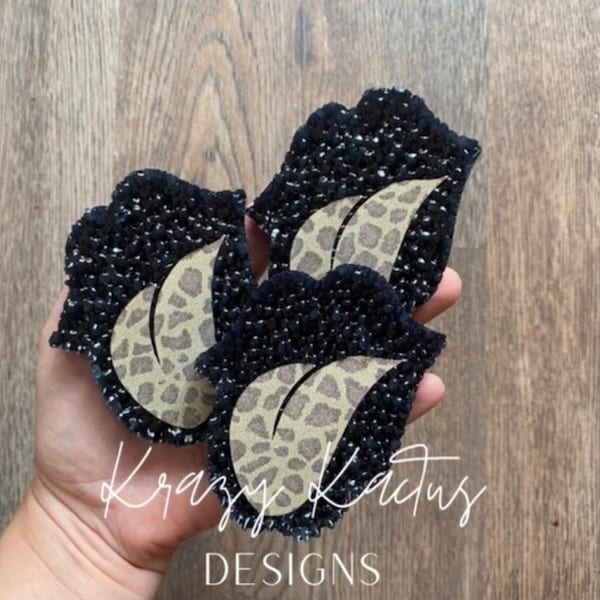 May include: Three black and white lips with a leopard print design. The lips are made of fabric and have a beaded texture. The lips are arranged in a stack, with the top lip slightly overlapping the middle lip. The bottom lip is completely visible. The lips are held in a hand, and the background is a wooden surface.