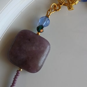 May include: A close-up of a purple square-shaped gemstone bead with a gold clasp and blue and green glass beads.