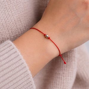 Op de afbeelding: Een rode draadarmband met een kleine zilveren kraal in het midden. De armband is om de pols geknoopt. De achtergrond is een persoon die een lichtbeige trui draagt.