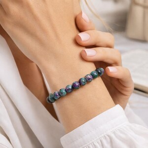 Peut inclure: Un bracelet de perles composé de pierres précieuses rondes et polies. Les perles sont un mélange de teintes vert foncé et violet. Le bracelet est porté au poignet, mettant en valeur les couleurs et le design.