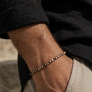 May include: A beaded bracelet with alternating brown and white beads. The bracelet is worn on a wrist, with a black shirt and tan pants in the background. The bracelet appears to be made of natural materials.
