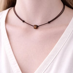 Könnte beinhalten: Eine braune Choker-Halskette mit einem kleinen, quadratischen Anhänger aus Tigeraugenstein. Die Halskette besteht aus zwei dünnen, dunkelbraunen Kordeln, die auf jeder Seite des Steins verknotet sind. Die Halskette wird über einem weißen Hemd getragen.