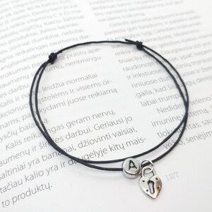 Puede incluir: Un brazalete de cuerda negro con un dije de plata en forma de corazón con un candado y la letra "A". El dije cuelga del brazalete.
