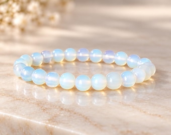 Könnte beinhalten: Ein elastisches Armband aus glatten, runden, durchscheinenden weißen Perlen mit einem dezenten irisierenden blauen Schimmer. Die Perlen sind von einheitlicher Größe und in einer durchgehenden kreisförmigen Schlaufe angeordnet.