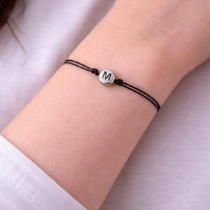Può includere: Braccialetto con cordino nero e ciondolo circolare color argento con la lettera "M". Il braccialetto è indossato su un polso, con una camicia bianca visibile sullo sfondo. Il braccialetto è un design semplice e minimalista.