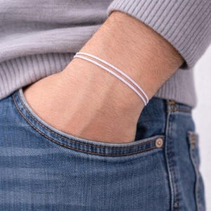 Puede incluir: Primer plano de una pulsera blanca con dos finas bandas. La pulsera se lleva en la muñeca. La persona lleva un jersey gris y vaqueros azules. La mano está en el bolsillo del vaquero.