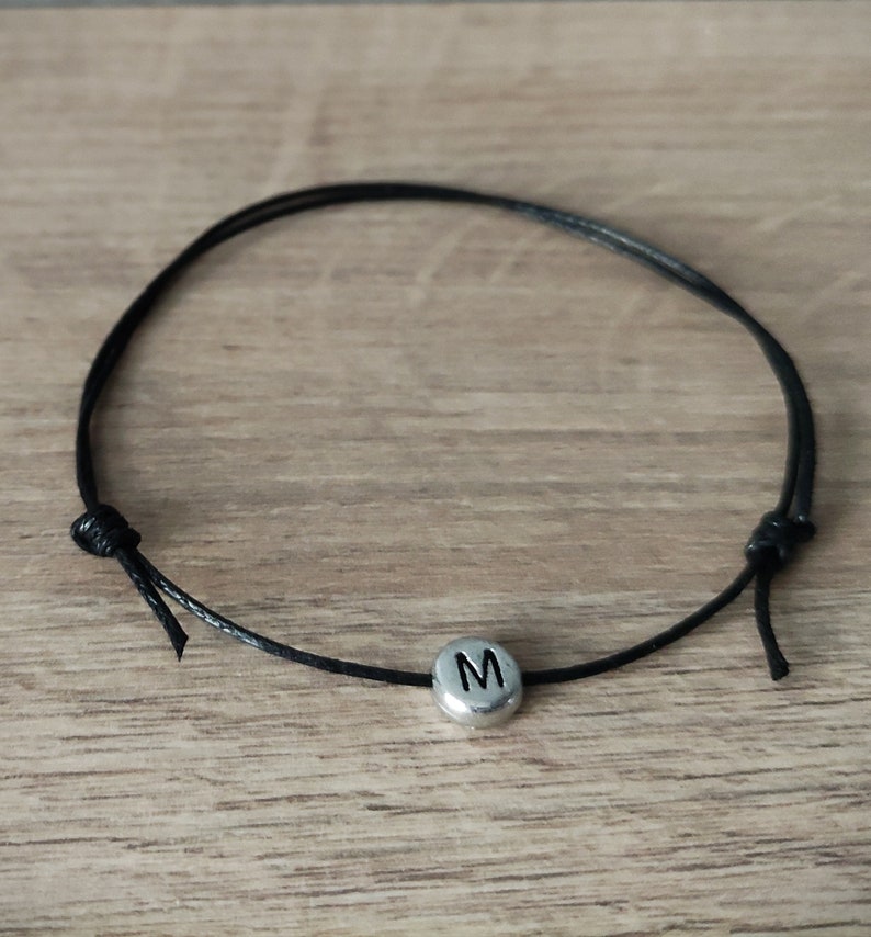 May include: A black cord bracelet with a silver-coloured bead featuring the letter "M". The bracelet is adjustable with sliding knots. The bracelet is displayed on a light brown wooden surface.