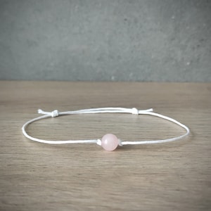 Puede incluir: Un sencillo brazalete de cuerda blanca con una sola cuenta de piedra rosa.