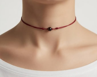 May include: A burgundy choker necklace featuring a dark, round bead as a focal point. The necklace is made of thin, burgundy-colored cord, knotted on each side of the bead. The necklace is worn around the neck.