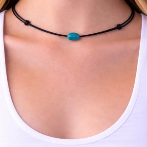 May include: A black choker necklace featuring a vibrant turquoise oval bead. The necklace is made of a thin, black cord and is worn around the neck. The design is simple and elegant, suitable for everyday wear.