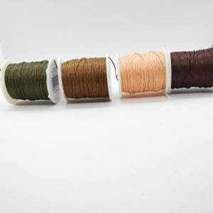 May include: Four spools of waxed cord in shades of olive green, light brown, peach, and dark brown. The cord is tightly wound on white plastic spools, suitable for crafts and jewellery making. The spools are arranged horizontally against a white background.