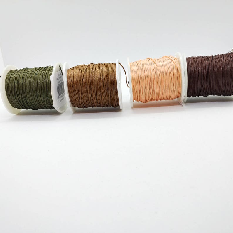 May include: Four spools of waxed cord in shades of olive green, light brown, peach, and dark brown. The cord is tightly wound on white plastic spools, suitable for crafts and jewellery making. The spools are arranged horizontally against a white background.