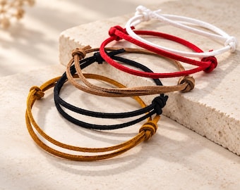 May include: A collection of four adjustable cord bracelets in white, red, brown, and black. Each bracelet features a thin, round cord with a sliding knot mechanism for closure, allowing the size to be adjusted. The cords have a matte, textured finish.