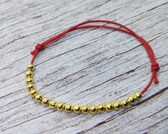 May include: A red string bracelet with gold beads. The bracelet is tied with a knot at the end.