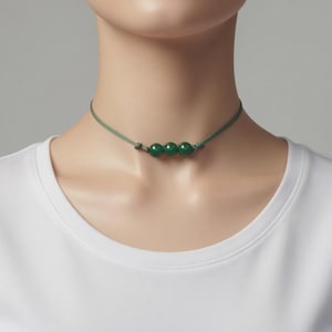 Op de afbeelding: Een groene choker-ketting met kralen. De ketting heeft drie ronde, donkergroene kralen geregen op een dun, lichtgroen koord. Het koord is aan weerszijden van de kralen geknoopt. De ketting wordt om de nek gedragen.