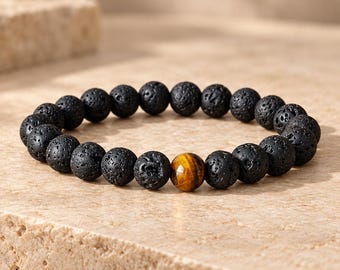 Puede incluir: Una pulsera de cuentas compuesta por una serie de piedras de lava negras, redondas y porosas, dispuestas en un patrón circular, acentuada por una cuenta de ojo de tigre marrón dorado, lisa y facetada, situada cerca del centro.