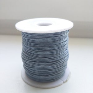 May include: A spool of grey waxed cord, suitable for jewellery making and crafts. The cord is tightly wound around a white plastic spool. The colour is a muted grey-blue, suitable for various projects.
