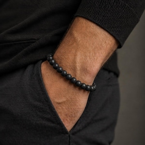Può includere: Braccialetto con perline in pietra lavica nera. Il braccialetto è composto da piccole perline rotonde e scure infilate insieme. Il braccialetto è indossato al polso, con la mano in tasca. La persona indossa una maglia nera a maniche lunghe.