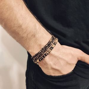 May include: A set of three bracelets made of brown and gold wooden beads. The bracelets are stacked on a person's wrist.