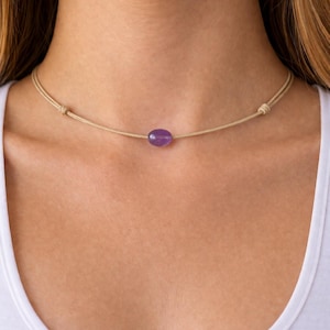 May include: A minimalist choker necklace featuring a small, oval-shaped, purple gemstone pendant. The pendant is suspended from a thin, beige cord that ties at the back. The necklace is simple and elegant, suitable for everyday wear.