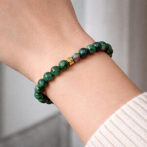 Puede incluir: Pulsera de cuentas de piedras preciosas verdes y rojas con una cuenta espaciadora dorada. La pulsera se lleva en la muñeca. Se ve un suéter color crema en el fondo. Las cuentas tienen aproximadamente 0,8 cm de diámetro.