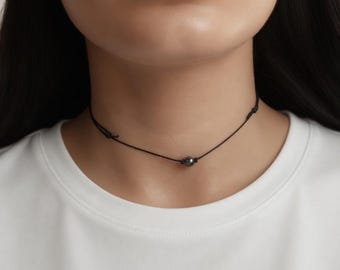 May include: A black choker necklace with a small, dark gray bead at the center. The necklace is worn around the neck, and the person is wearing a white t-shirt. The background is a neutral color.