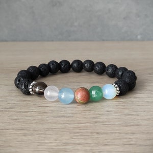 May include: A black lava rock bracelet with six gemstone beads. The beads are a dark brown, light pink, light blue, red, green, and white.
