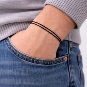 Puede incluir: Primer plano de una muñeca adornada con dos pulseras finas y negras. La persona lleva un jersey gris y vaqueros azules. Las pulseras son de diseño sencillo y minimalista, adecuadas para el uso diario. Cada pulsera tiene unos 0,2 cm de grosor.