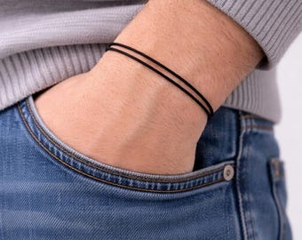 May include: A close-up of a wrist adorned with two thin, black bracelets. The person is wearing a gray sweater and blue jeans. The bracelets are simple and minimalist, suitable for everyday wear.