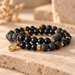 Op de afbeelding: Een dubbelstrengs kralenarmband met afwisselend zwarte lavasteenkralen en gladde zwarte onyxkralen. De armband bevat goudkleurige metalen tussenstukjes met ingewikkelde patronen en een goudkleurige Hamsa-hand bedel.