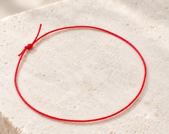Könnte beinhalten: Ein dünnes, leuchtend rotes Schnurarmband in kreisförmiger Anordnung. Das Armband weist an einer Seite ein einfaches Knotendetail auf, von dem zwei kurze, lose Enden abstehen, was ein minimalistisches Design aus feinem Kordelmaterial zeigt.