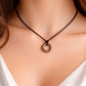 Może przedstawiać: Minimalistyczny naszyjnik z małą, okrągłą zawieszką w stonowanym odcieniu oliwkowej zieleni. Zawieszka zawieszona jest na delikatnym, czarnym sznurku, tworząc prosty, ale elegancki design. Naszyjnik prezentowany jest na neutralnym tle.