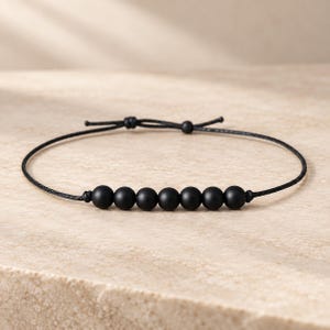 Könnte beinhalten: Ein schwarzes Kordelarmband mit sieben mattschwarzen, kugelförmigen Perlen in einer Reihe. Das Armband verfügt über einen verstellbaren Schiebeknotenverschluss, bei dem zwei dünne schwarze Kordelenden aus dem mittleren Knoten für einen sicheren Sitz.