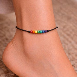 Puede incluir: Una tobillera negra con una fila de cuentas de colores del arcoíris. Las cuentas son rojas, naranjas, amarillas, verdes, azules y moradas. La tobillera se lleva en el tobillo.