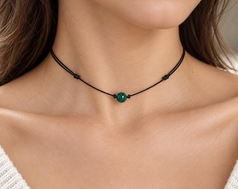 May include: A choker necklace featuring a single round malachite bead with distinct green and black banded patterns. The bead is centered on a thin black cord, which is secured by two small knots on either side of the stone to keep it in place.