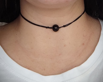 May include: A black choker necklace featuring a dark, round bead as a focal point. The bead is centered on a thin, dark cord, creating a minimalist design. The necklace is worn around the neck.