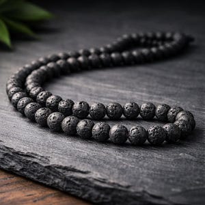 Puede incluir: Un collar de cuentas de piedra de lava negra. Las cuentas redondas tienen una textura porosa. El collar se muestra sobre una superficie gris oscuro. Las cuentas son de color gris oscuro.